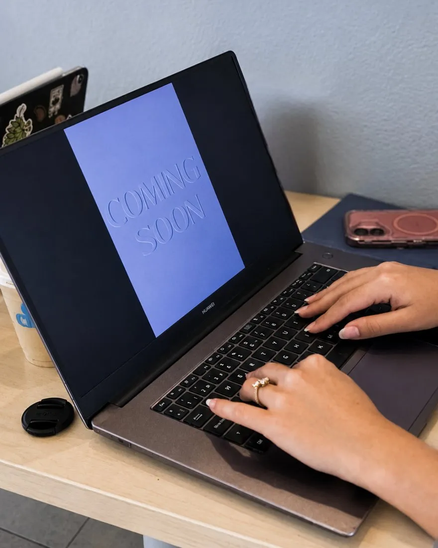 Manos trabajando en una laptop con el diseño de una publicación “Coming soon” en pantalla